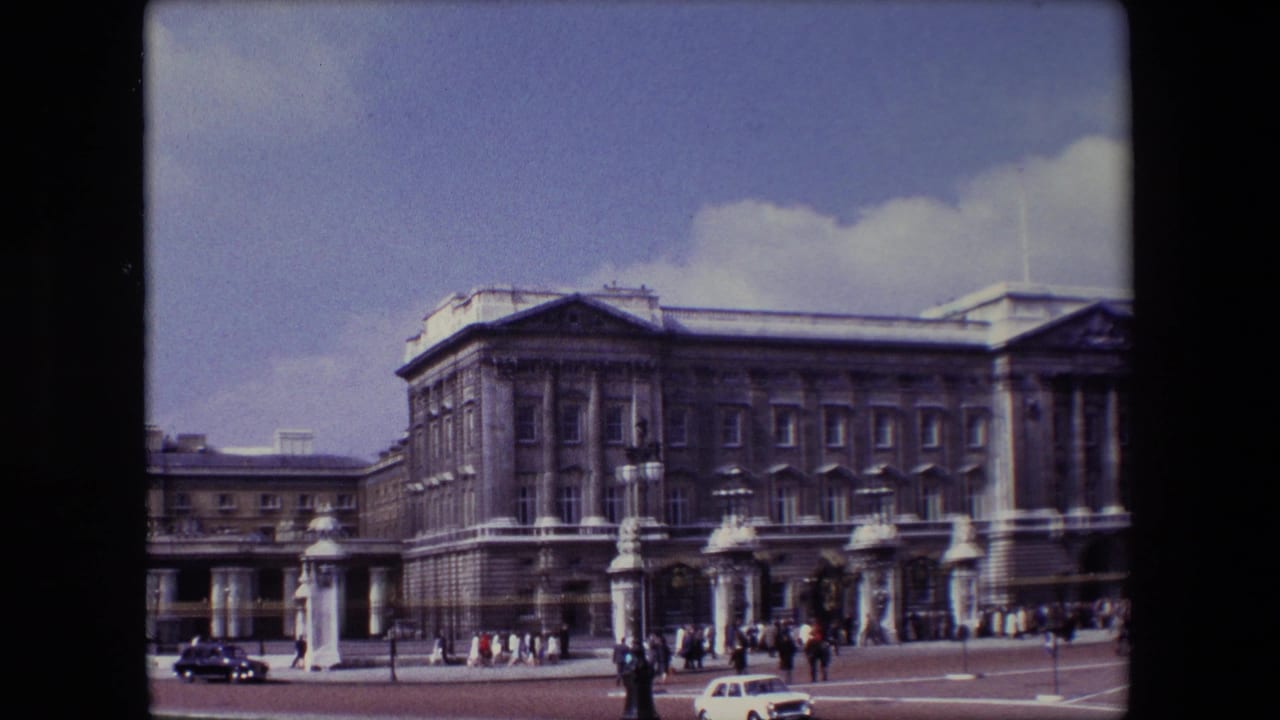 Wide view of Buckingham Palace with pedestrians, gates, and cars in the foreground.
