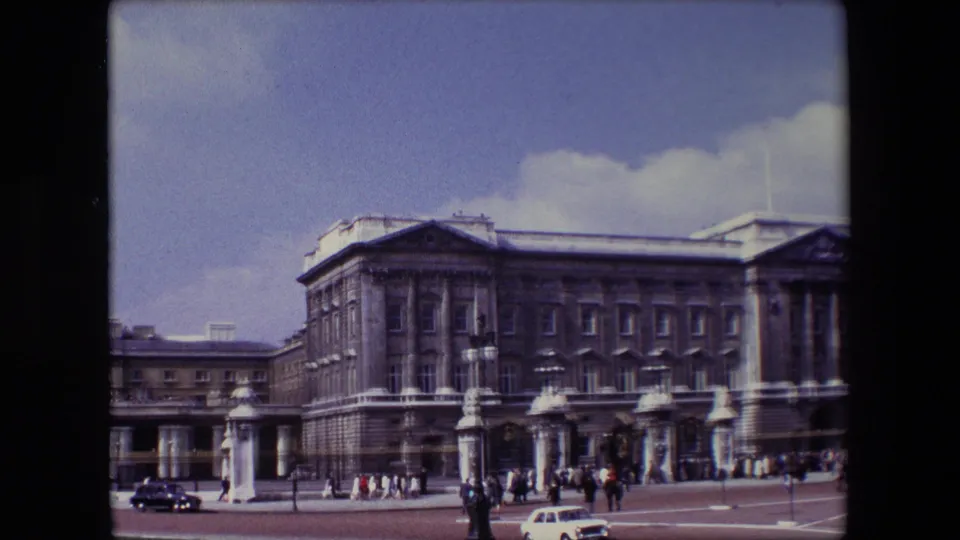 1967 8mm home-movie frame showing Buckingham Palace in London with tourists near the gates and classic cars passing across the foreground.