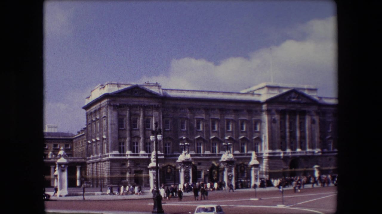 Buckingham Palace facade with people gathered near the gates and a small car at the lower edge.