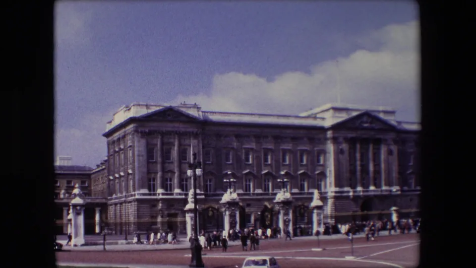 Wide 1967 London frame of Buckingham Palace, showing the neoclassical facade, forecourt gates, clustered visitors, and a small car near the bottom of the view.