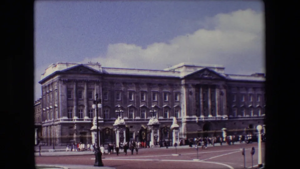 1967 8mm frame of Buckingham Palace in London, with tourists and pedestrians along the forecourt, a prominent lamppost, and pale road markings in front.