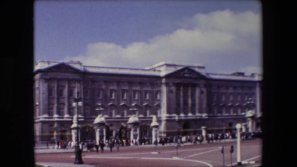 Final 1967 London frame holding on Buckingham Palace with visitors near the gates, roadway markings in front, and a white roadside post at the right edge.