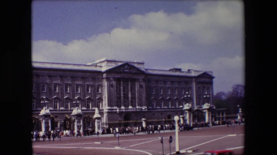 A 1967 home-movie frame shows the Buckingham Palace facade cropped closer, with visitors clustered near the gates and a red vehicle edge partly visible at the bottom.