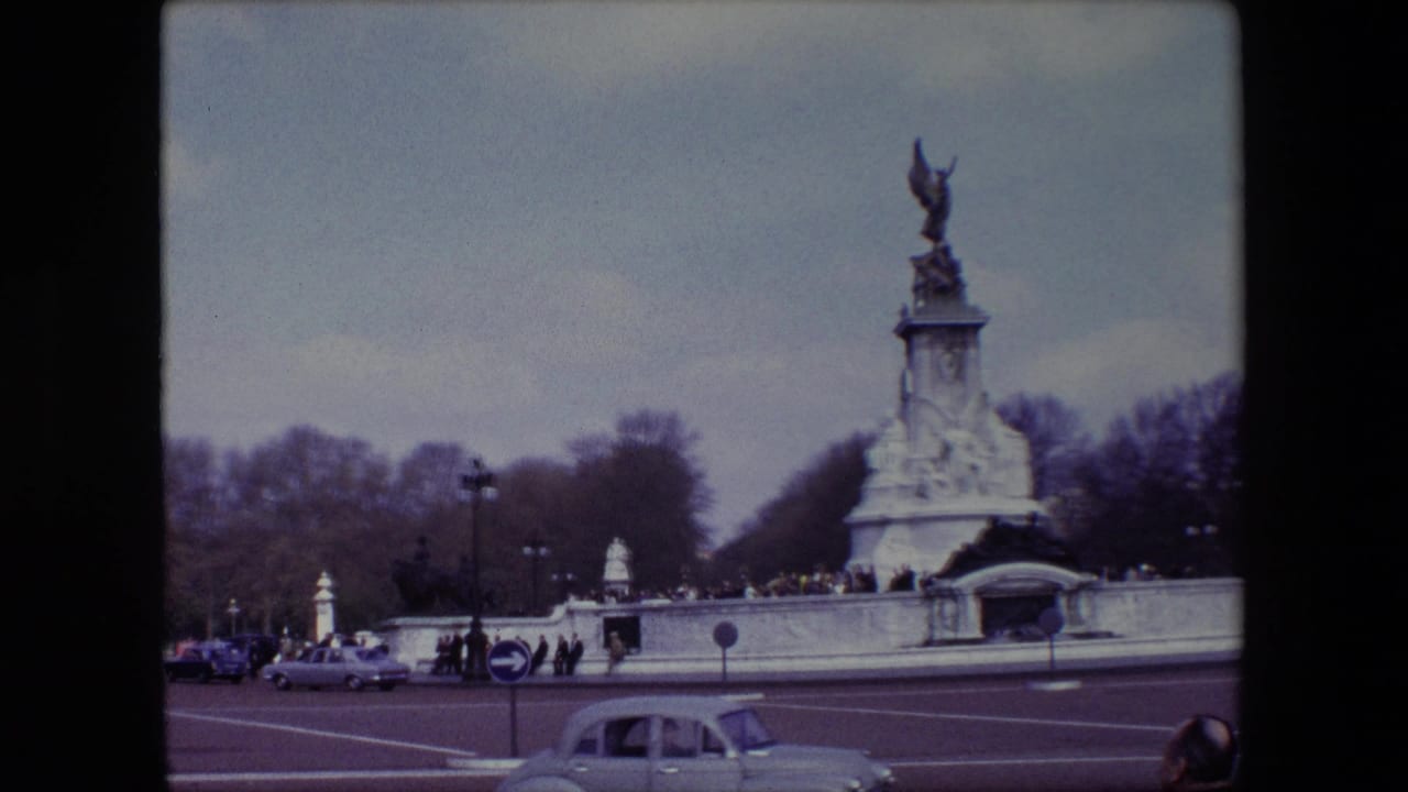 1967 London home-movie frame showing the Victoria Memorial at Buckingham Palace across the road, with classic cars, pedestrians, bare trees, and an overcast sky.