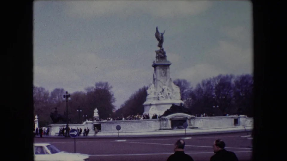 1967 London frame of the Victoria Memorial and Buckingham Palace forecourt wall, with pedestrians near the monument and two foreground figures watching from across the road.