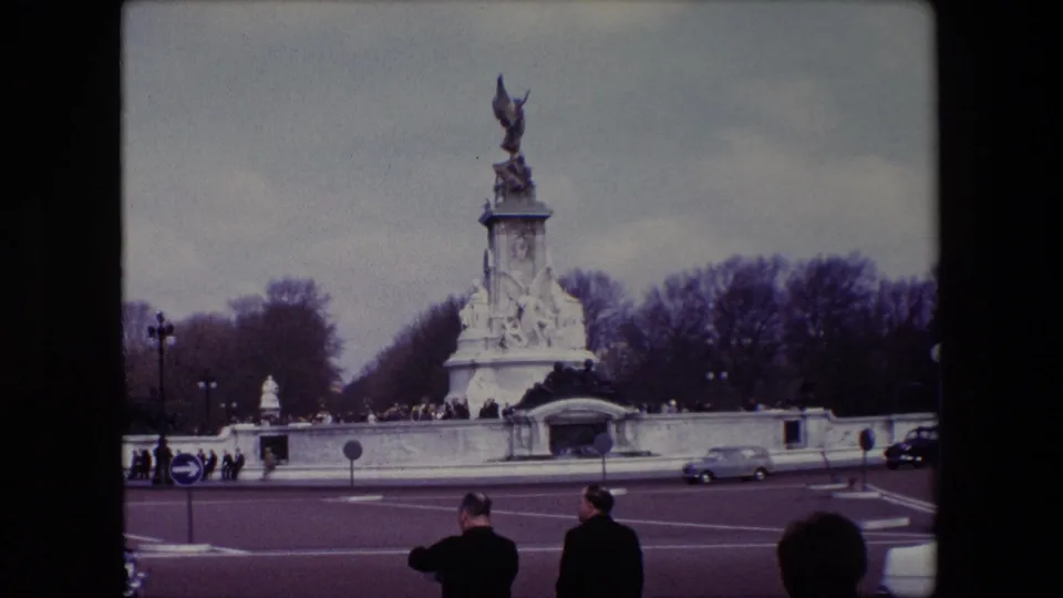 1967 London home-movie view of the Victoria Memorial across the roadway, with classic cars passing, pedestrians in coats, and bare trees behind the monument.