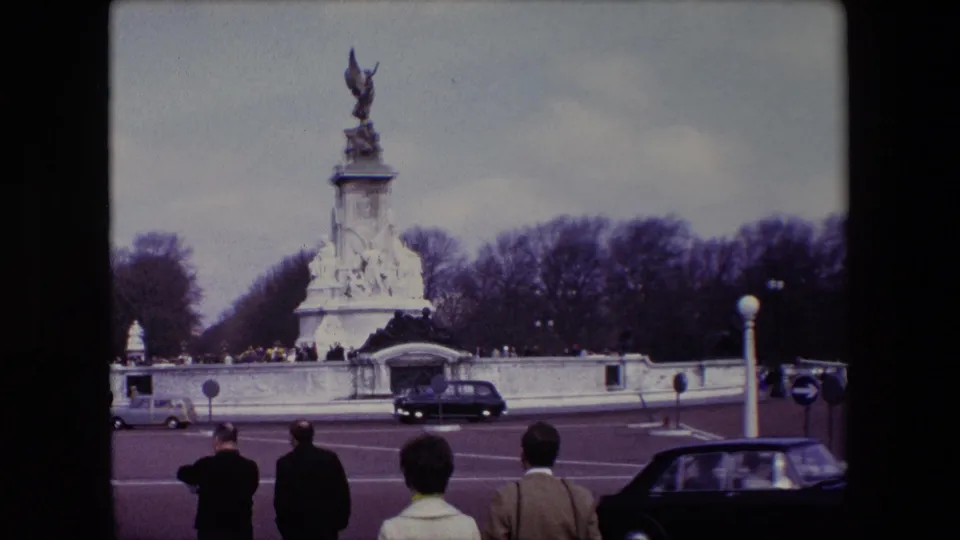 1967 London frame showing the Victoria Memorial at Buckingham Palace, foreground pedestrians seen from behind, and period cars moving through the traffic circle.