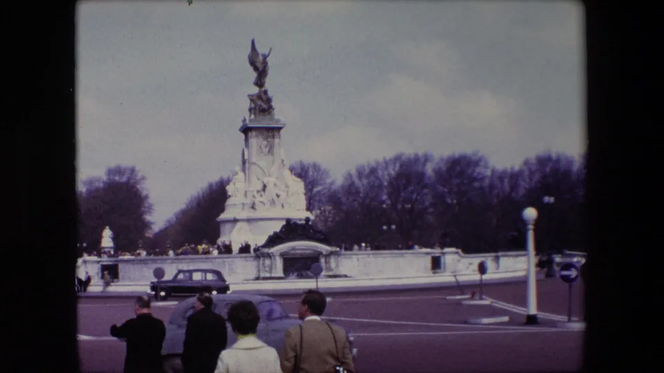 Wide 1967 London home-movie view of the Victoria Memorial across the road, with pedestrians in coats and a classic car passing in front.