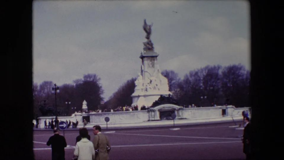 Wide home-movie frame of the Victoria Memorial in London, showing the monument, roadway, road signs, bare trees, and pedestrians in period coats.