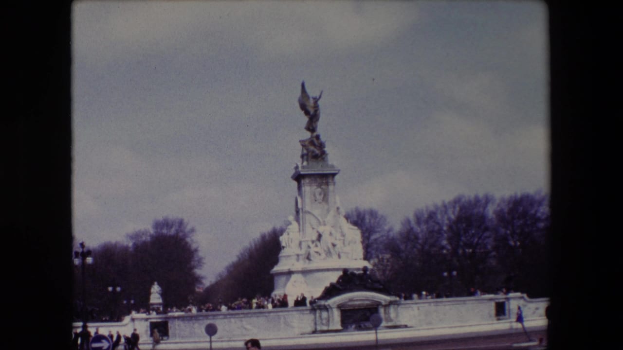 1967 London home-movie frame showing the Victoria Memorial in front of Buckingham Palace, with distant visitors near the low wall and bare trees beneath an overcast sky.