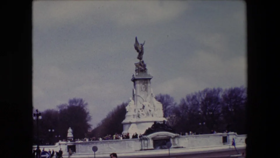 1967 London home-movie frame showing the Victoria Memorial in front of Buckingham Palace, with distant visitors near the low wall and bare trees beneath an overcast sky.