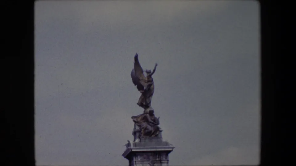 Closer 1967 London home-movie view of the Victoria Memorial's winged bronze figure, with the statue rising above the marble base against a pale overcast sky.
