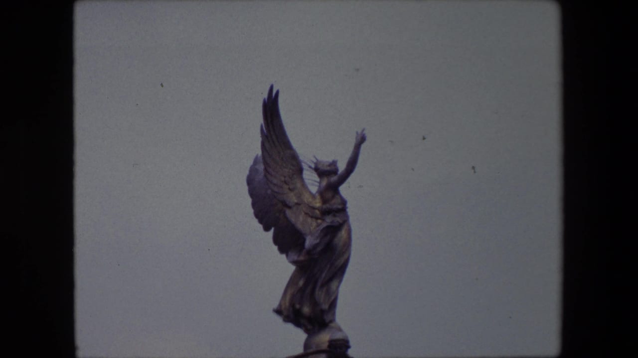 1967 London home-movie frame showing a bronze-toned winged angel statue reaching upward against a pale blue sky.