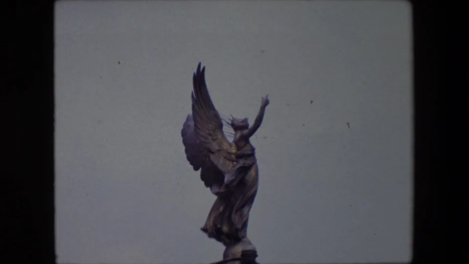 1967 London home-movie frame showing a bronze-toned winged angel statue reaching upward against a pale blue sky.