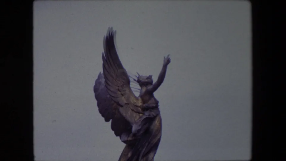 1967 London 8mm frame with the angel sculpture cropped closer, emphasizing the raised wing, arm, and draped figure against open sky.