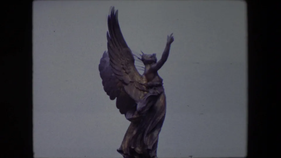 1967 London home-movie frame showing the winged statue from a slightly lower crop, with the figure and robe visible against the sky.