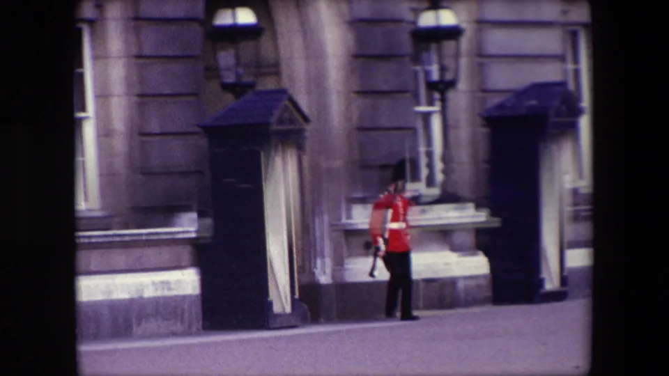 1967 London 8mm frame showing a British royal guard in a red tunic near a black sentry box, stone facade, sash window, and ornate lamppost.
