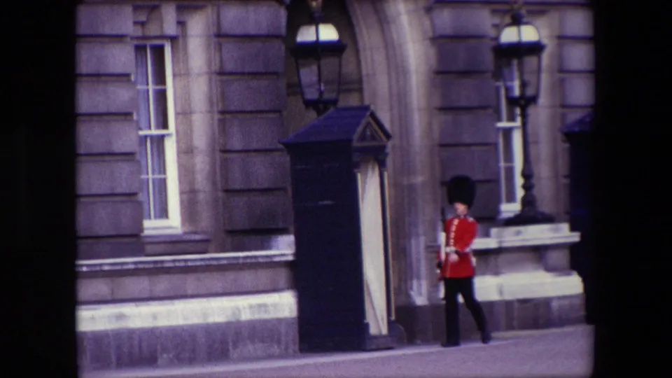 1967 London 8mm frame of a British royal guard in red uniform and black hat marching near a sentry box outside a formal stone building.
