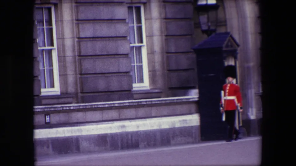 1967 London 8mm frame showing the back of a British royal guard in red tunic as he moves beside a black sentry box and stone building wall.