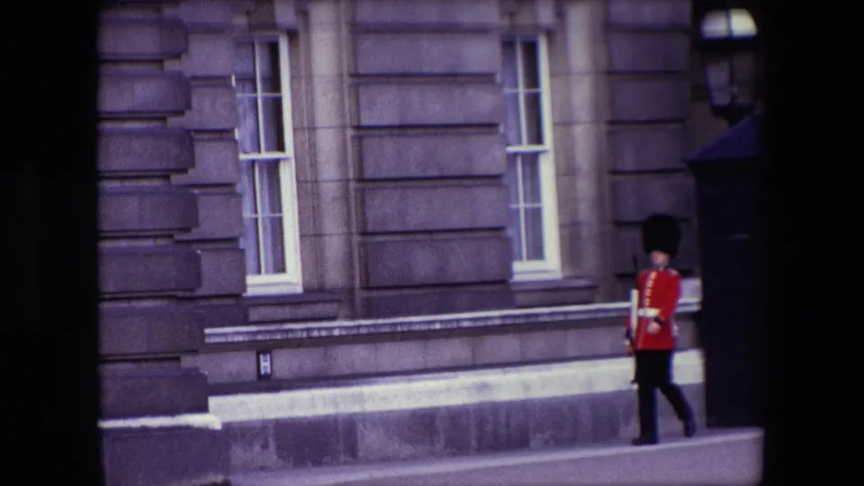 1967 London 8mm frame of a British royal guard walking along the paved forecourt in front of a grand stone facade with tall sash windows.