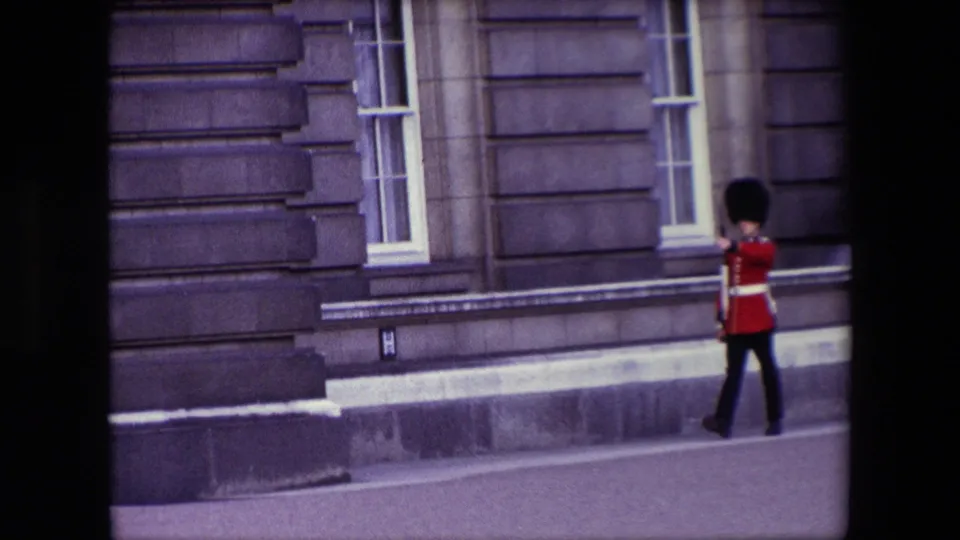 1967 London home-movie frame showing a British guard in a red ceremonial coat and tall black hat walking beside a grand stone building.