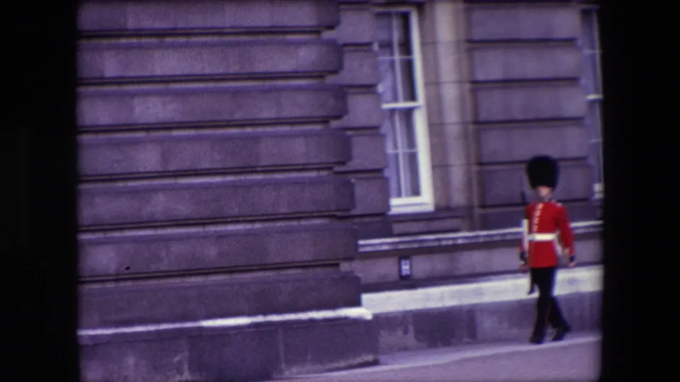1967 London frame of a British ceremonial guard in red coat and tall black hat moving past stonework and narrow windows.