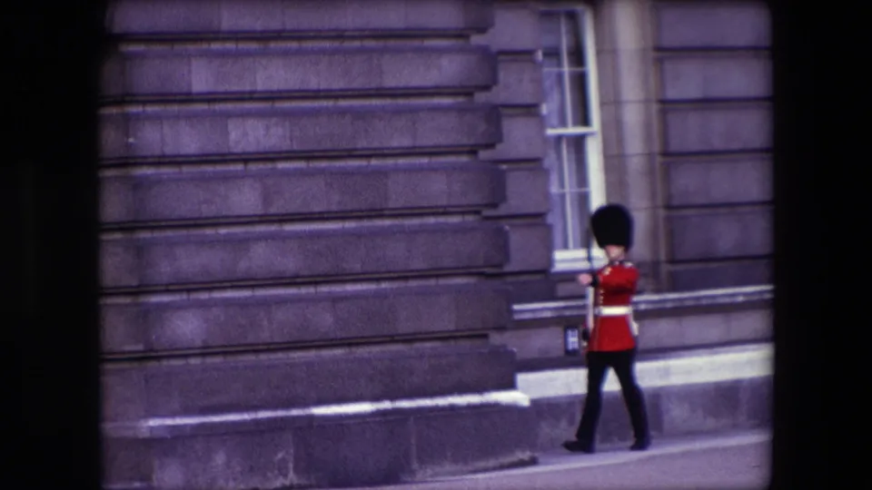 A British guard in red ceremonial uniform marches across the lower part of the frame against a London stone building in 1967 8mm footage.