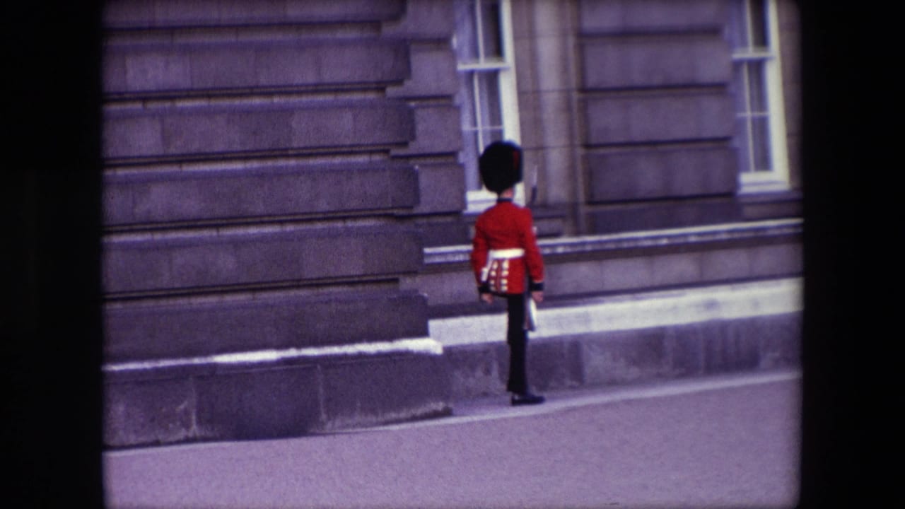 British Royal Guard in a red tunic standing beside a stone building wall.