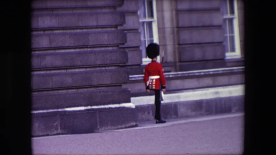 A British Royal Guard in red ceremonial uniform stands near a stone facade with tall windows in London, supported by the 1967 clip metadata.