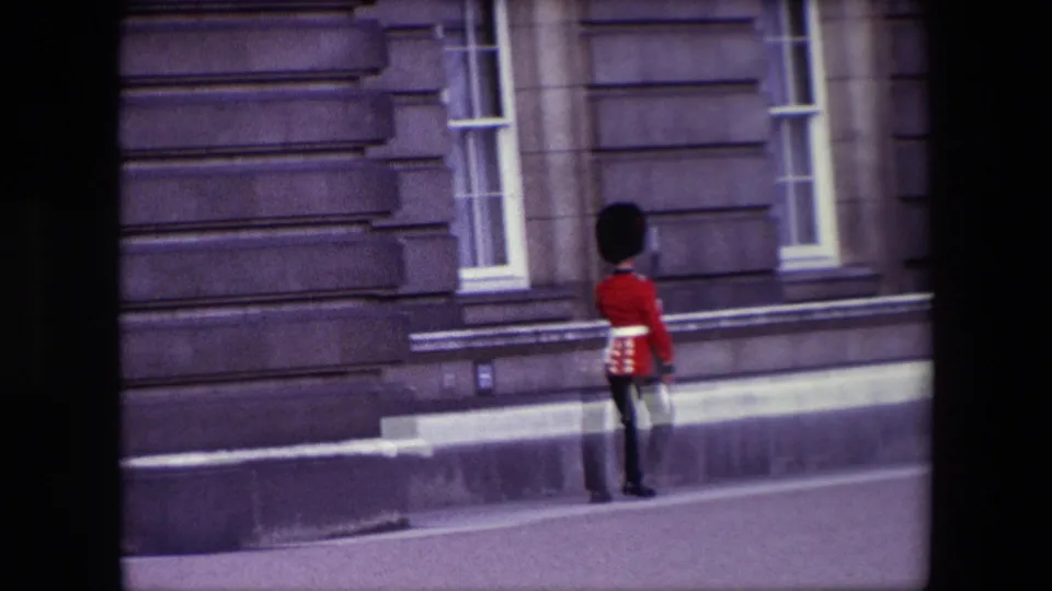 The guard moves along the pavement in front of formal stone architecture and tall windows, matching the London ceremonial context in the metadata.