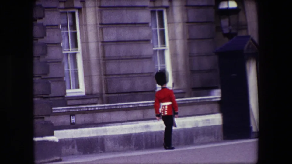 A British Royal Guard in red uniform stands or marches near a dark sentry box against a stone building in the supplied London 1967 setting.