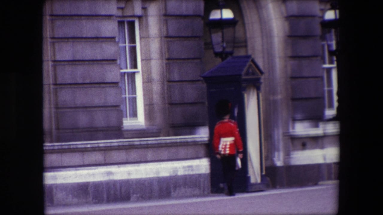 Guard in red tunic stands near a dark sentry box outside a stone building.