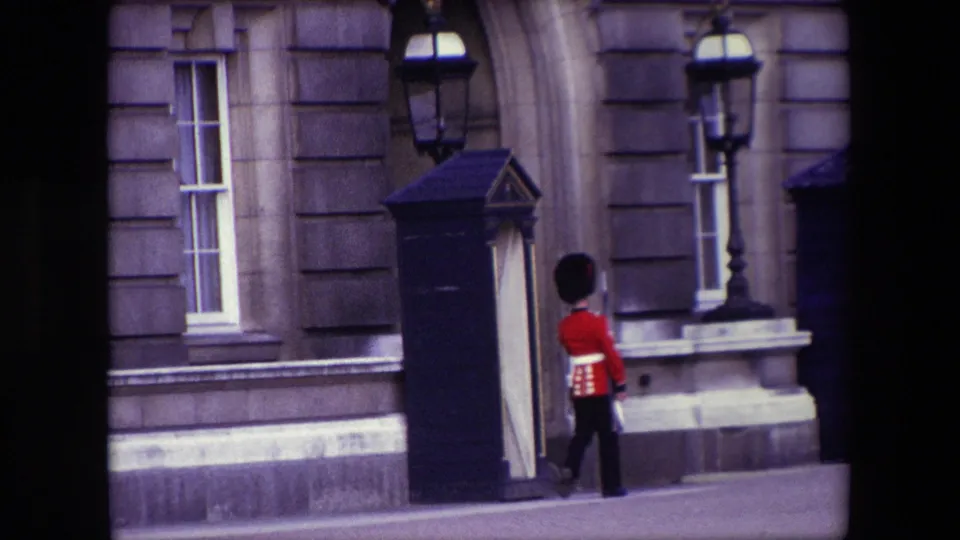 The uniformed guard moves beside the sentry box outside a formal stone building in London, with tall windows and lamps visible behind him.