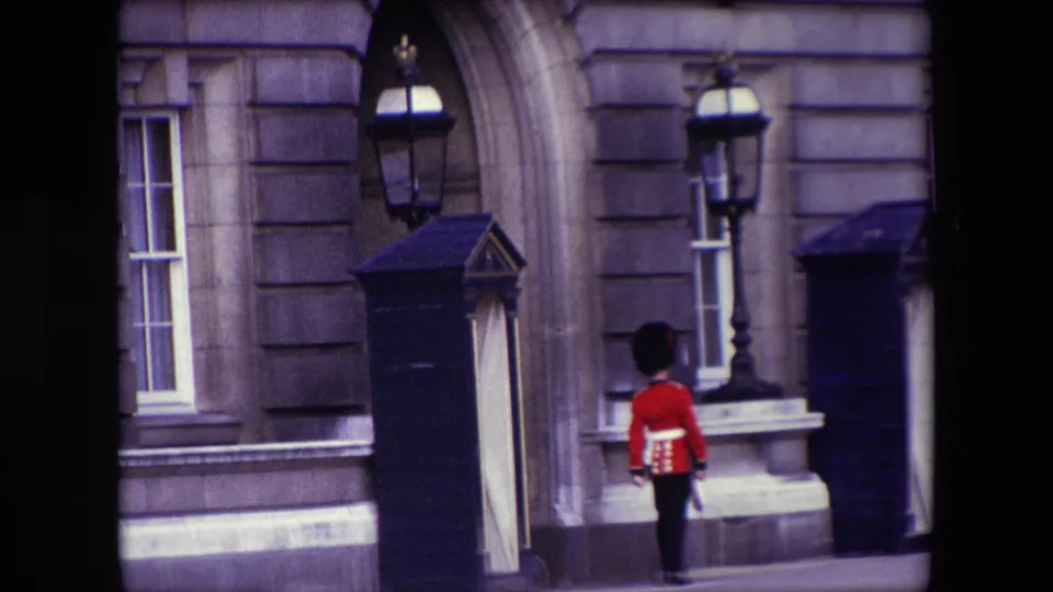 The guard is viewed from behind in 1967 London footage, positioned near a sentry box, arched stone doorway, and building-mounted lamp.