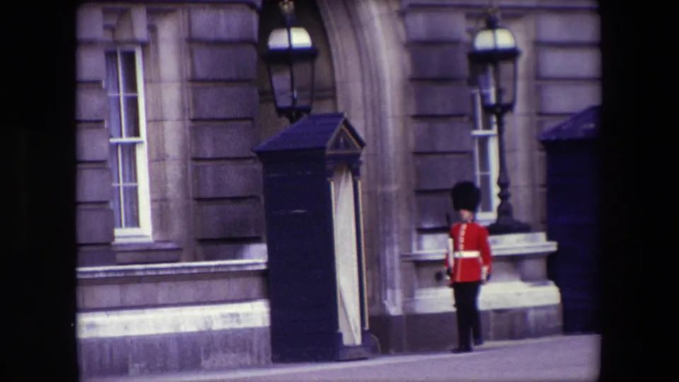 A Queen's Guard-style sentry stands facing the building near a dark sentry box, framed by stone architecture and a wall-mounted lamp in London.