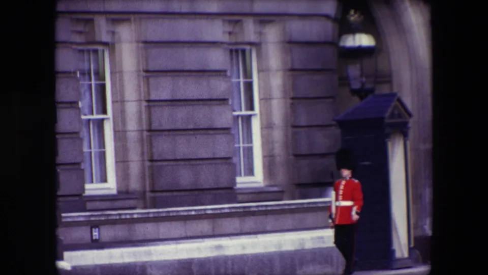1967 London home-movie frame with the Royal Guard moving along the walkway beside a sentry box and formal palace-style stonework.