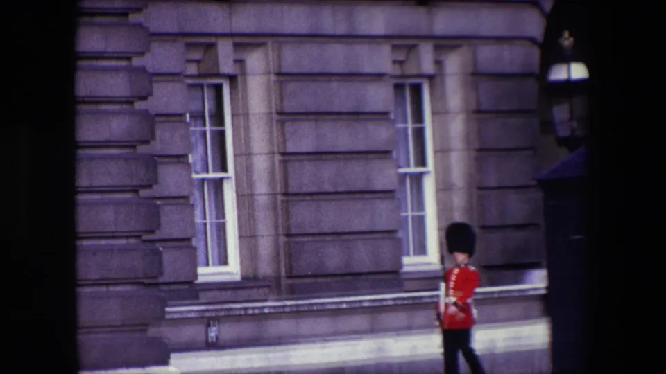 1967 London frame showing the guard partly lower in the composition as he walks past tall windows, carved stonework, and the edge of a sentry box.