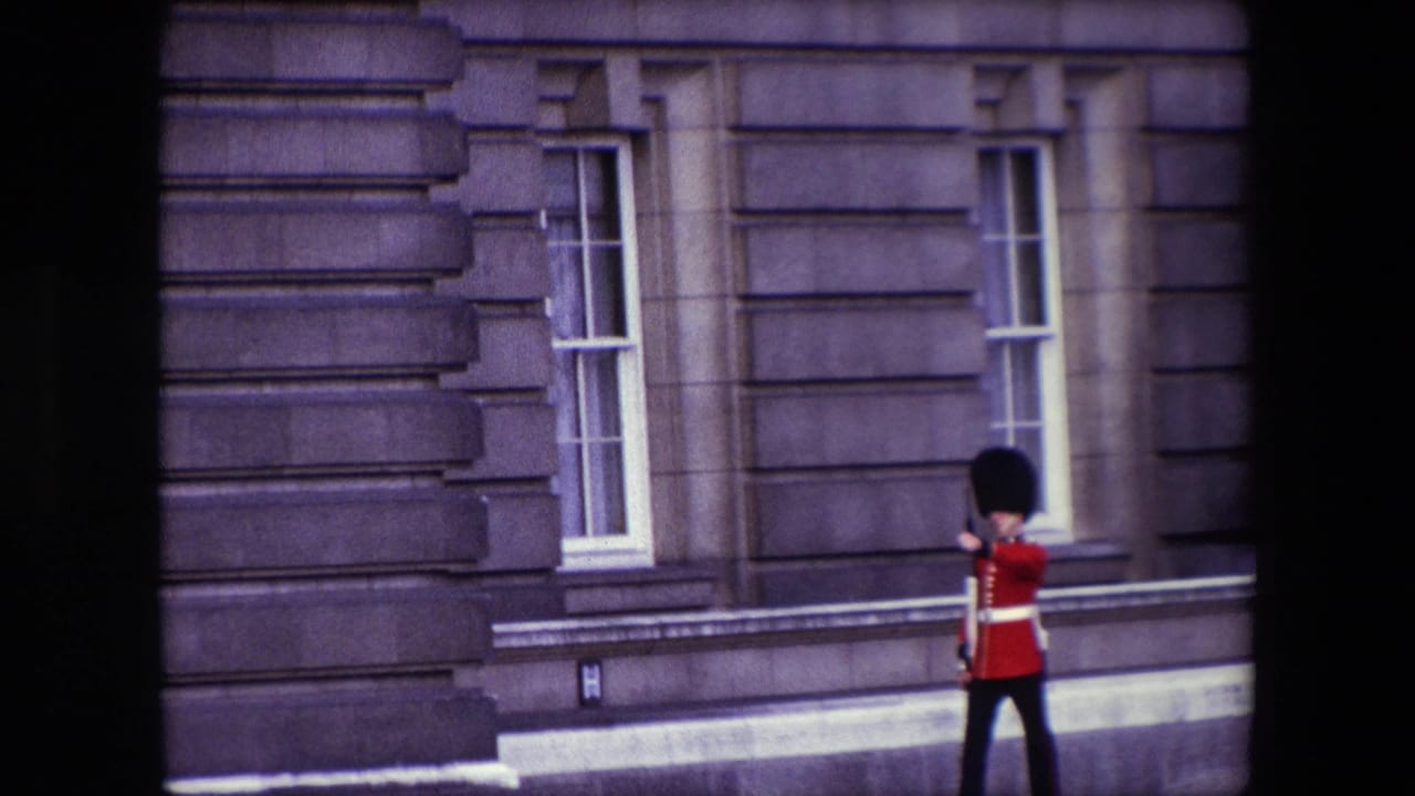 Queen's Guard marches past stone palace windows in a red tunic and tall black hat.