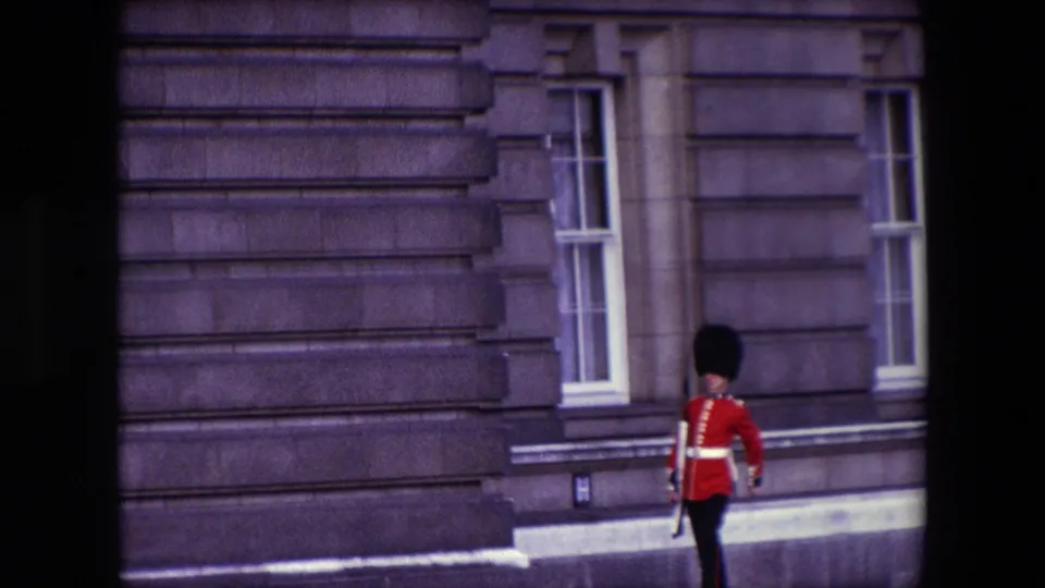 1967 London frame of a ceremonial guard moving in profile along a stone palace wall, with dark home-movie framing at the edges.