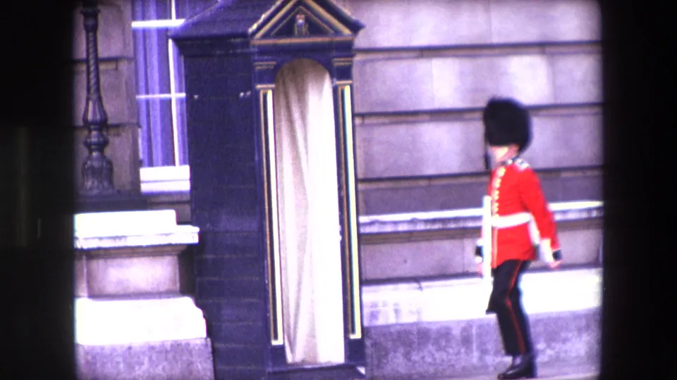1967 London home-movie frame showing the Queen's Guard walking near a black sentry box in front of a palace-style stone exterior.