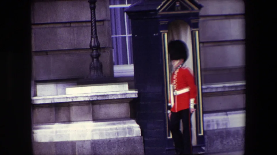 1967 London frame showing a Queen's Guard paused beside a black sentry box, with palace stonework and a window behind him.