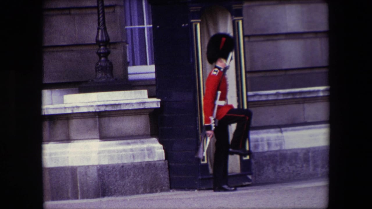 A British palace guard in red ceremonial uniform and tall black hat marches near a black sentry box outside a stone building in 1967 London home-movie footage.