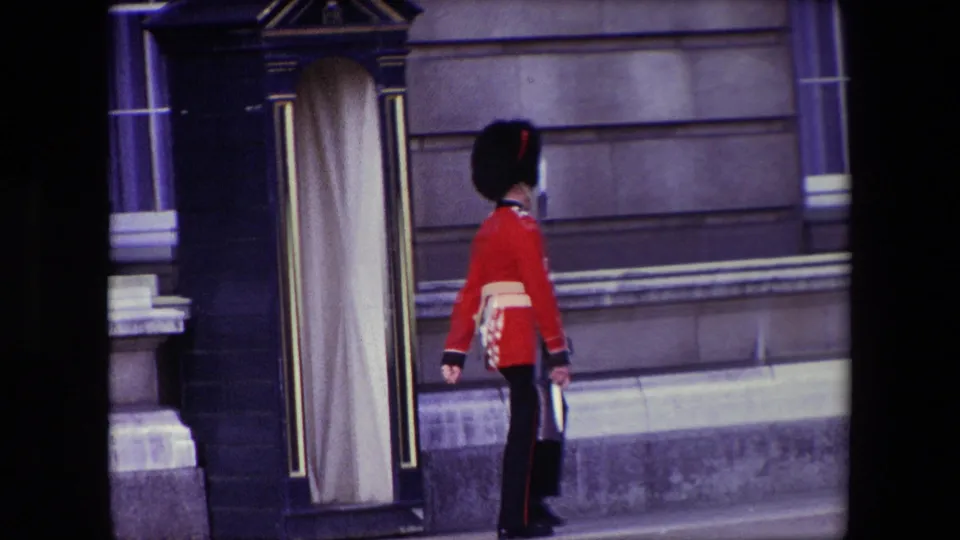 A ceremonial guard in red coat and tall black hat walks past a black sentry box and doorway against a formal London stone facade.