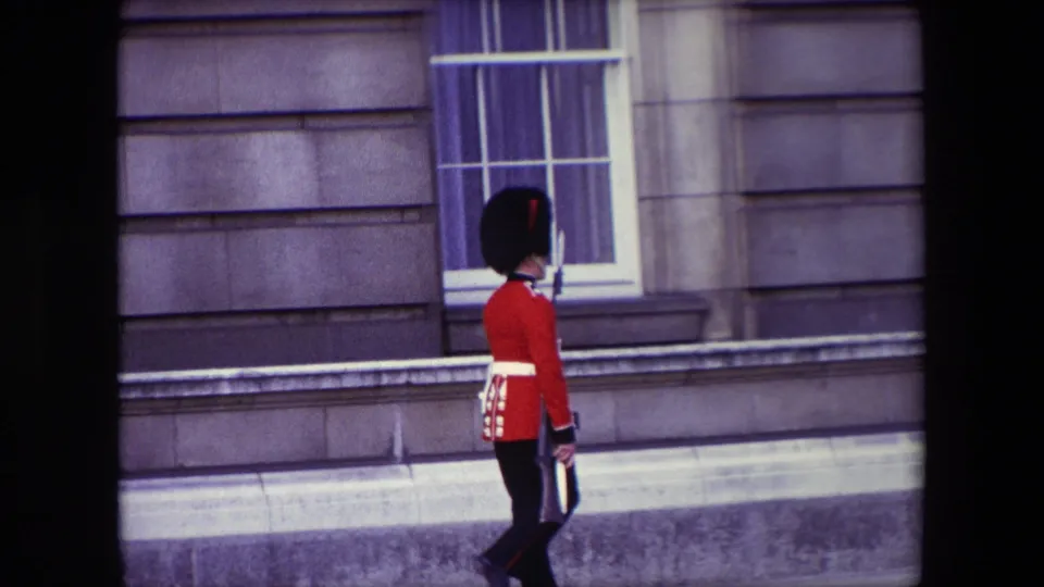 The guard continues marching along the stone facade below a large sash window, filmed in soft 8mm color from a visitor viewpoint.