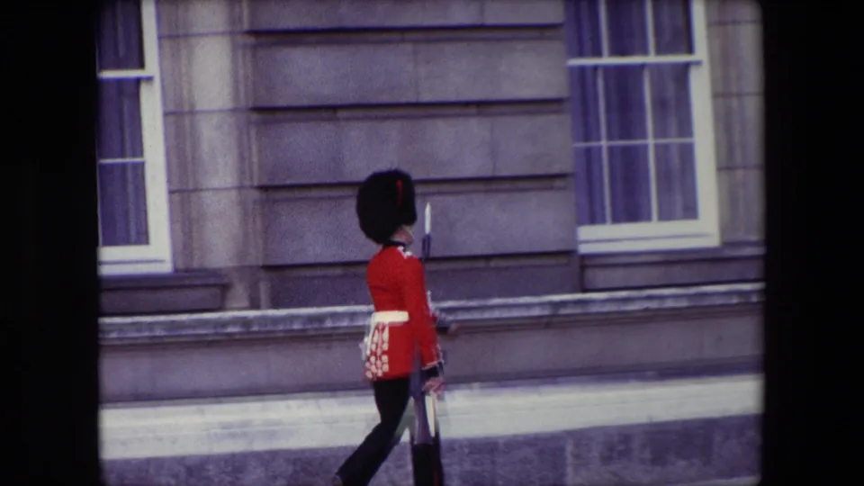 A British ceremonial guard in red uniform marches past a stone building facade and window in brief 1967 London 8mm footage.