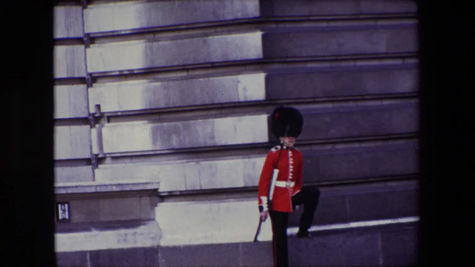 The Queen's Guard faces toward the camera area with one leg raised in a formal step beside the London stone facade in the 1967 home-movie footage.