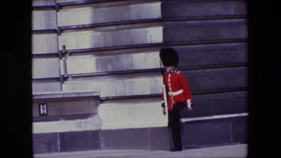 1967 London home-movie frame showing a Buckingham Palace guard in red ceremonial tunic and black bearskin-style hat near a pale stone facade.