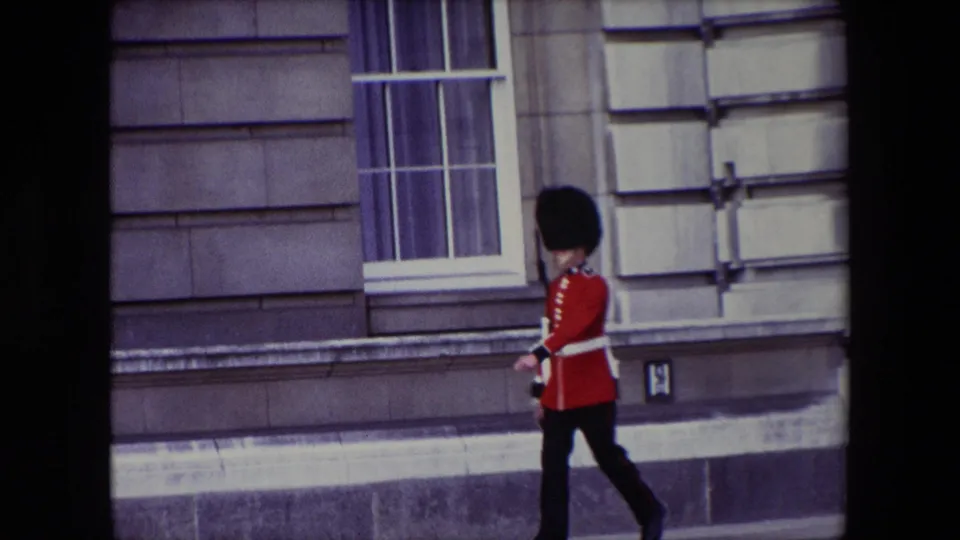 1967 London home-movie frame showing the guard in profile during the ceremonial movement, with a rifle and palace window behind him.