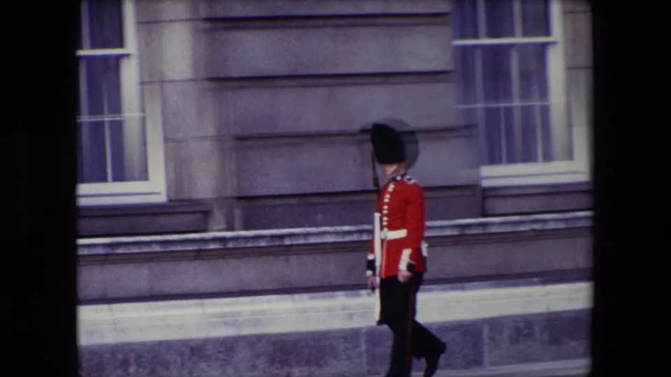 1967 London frame of a Buckingham Palace guard completing the movement along the pale stone exterior, with window frames and masonry visible behind him.