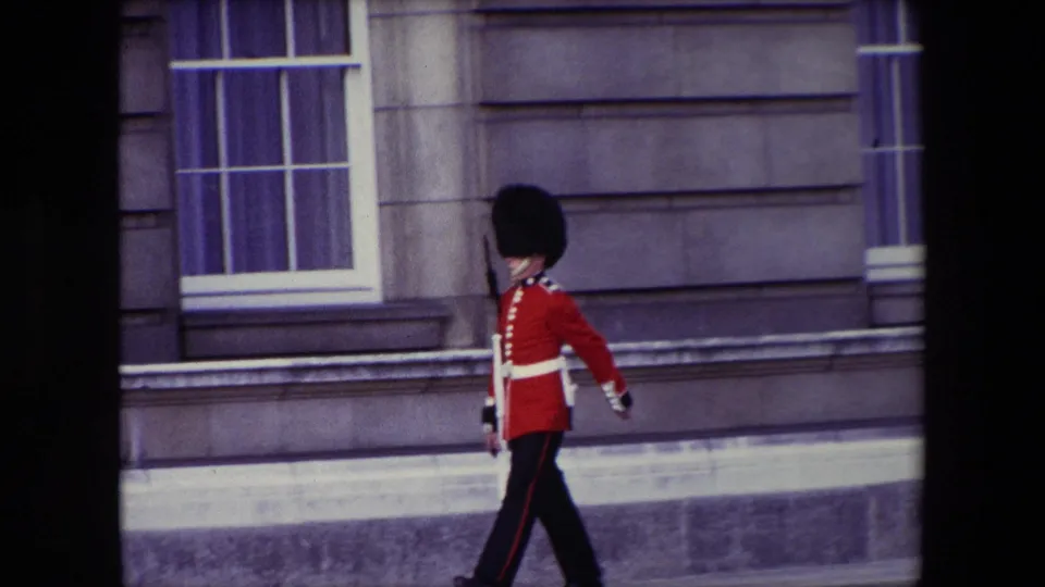 A Queen's Guard in red tunic and tall black hat marches along a pavement in London, England, supported by the supplied 1967 metadata.